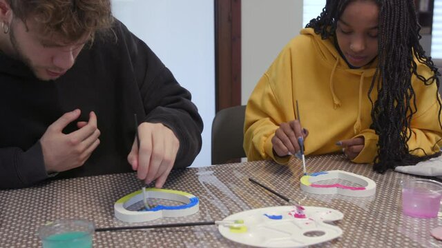 Boyfriend and girlfriend smiling while painting wooden hearts at home. Domestic life for interracial couple doing leisure activities with paint for fun. Happy people laughing on Valentine's Day
