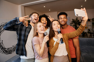 Selfie time. Group of young happy multicultural friends taking a selfie using smartphone while spending time together, playing karaoke at home