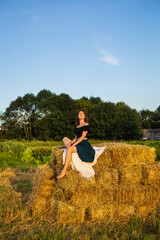 Naklejka na ściany i meble Young beautiful woman sits on a large pile of straw bales. Freedom, wind, setting sun.