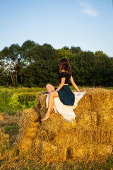 Young beautiful woman sits on a large pile of straw bales. Freedom, wind, setting sun.