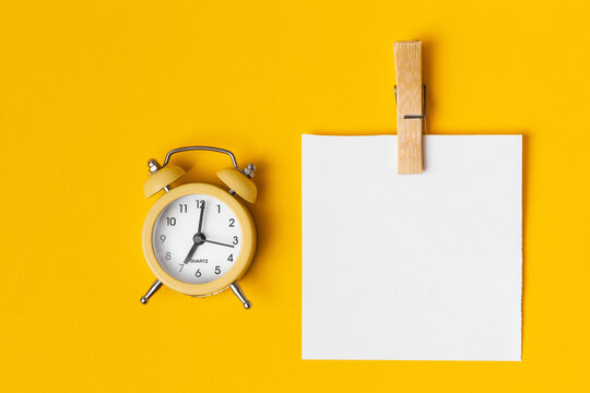Top view of the blank white paper for your notes to remember pinned to the yellow background with a wooden pin next to the yellow alarm clock