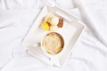 Cup of coffee, macaroons on the square tray in the middle on the white bedsheet. Service. Holiday. Morning surprise. Coffee break atmosphere. 