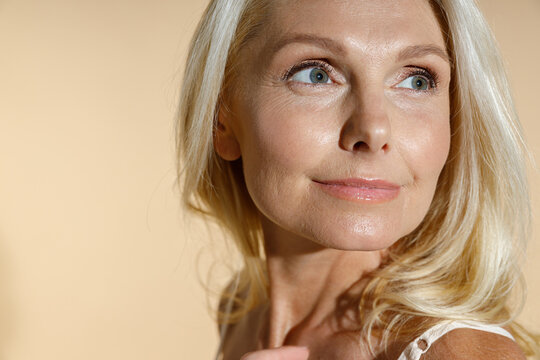 Close up portrait of adorable caucasian mature blonde woman with natural makeup looking aside while posing isolated over beige background