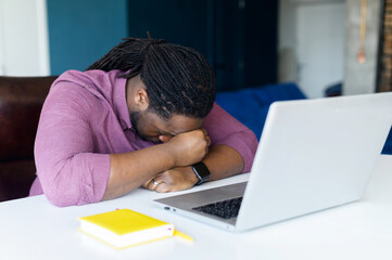 Exhausted African-American man with locks hair takes a nap on the workplace, tired freelancer guy sleeps right on the table in front of the laptop. Overworked and overload concept