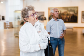 mature woman  examines paintings in an exhibition in hall of an art museum