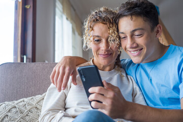 Smiling middle-aged 40s mother rest with grown-up son using smartphone together, happy young man...