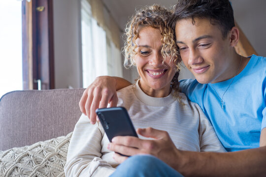 Smiling middle-aged 40s mother rest with grown-up son using smartphone together, happy young man enjoy family weekend with mom browsing wireless Internet on cellphone, have fun at home
