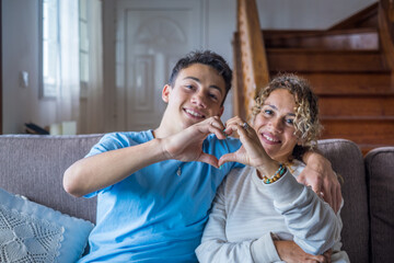 Portrait of couple of mom and son together a home making a heart love shape with their fingers and hands with the faces at the background. Affectionate teenager and woman enjoying