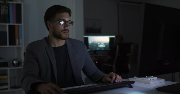 Lateral dolly of young caucasian handsome businessman in eye glasses late at night at office in front of computer. Male broker following rates and bets. Focused man working late.