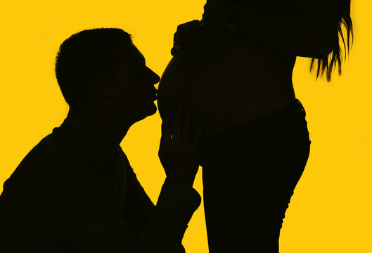 Enamored husband, man kisses the belly of a pregnant girl with long hair close-up on a yellow background silhouette. Pregnancy photo.