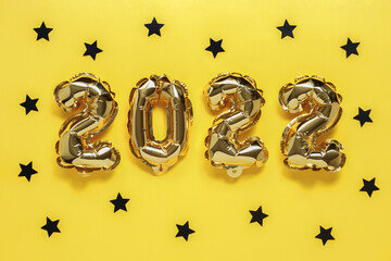 Fototapeta premium Christmas or New Year background. Text 2022, made of golden balloons, on a yellow background, with black confetti stars. Postcard or calendar. Merry Christmas or Happy New Year.