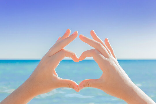 The hands of a young girl depict a sign or symbol of heart and love against the backdrop of the sea and the sky