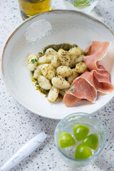 Plate of italian gnocchi with basil pesto sauce and prosciutto ham, vertical shot on a beige stone surface, selective focus