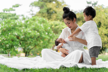 Asian young beautiful mother holding her newborn is sleeping and feel with love and touching gently then sitting on green grass in the park