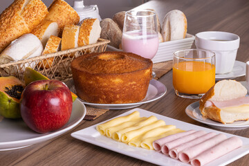 Mesa de café da manhã com vários alimentos, incluindo pães, doces, bolo, iogurte, chá, café, suco de laranja, presunto, queijo, mamão e maçã
