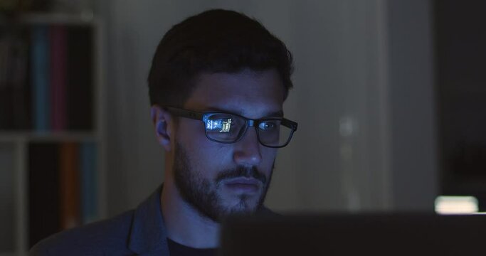 Lateral dolly of young caucasian handsome businessman in eye glasses late at night scrolling in front of computer. Financial animations reflected in glasses. Male broker following rates and bets.