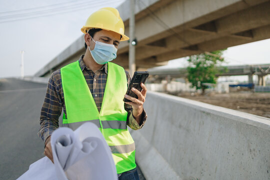 asian construction worker wearing protective masks with holding blueprint and using smartphone in construction site