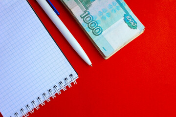 On a red desktop is a notebook, a pen and Russian money in denomination of 1000 one thousand rubles.