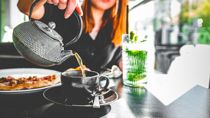 woman hand with metal teapot pour black tea in black glass cup in cafe