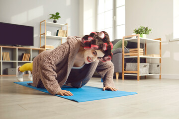 Young woman exercising on fitness mat and smiling. Funny housewife in hair curlers and beauty face mask doing push-ups during sports workout at home. Concept of people keeping body fit and healthy