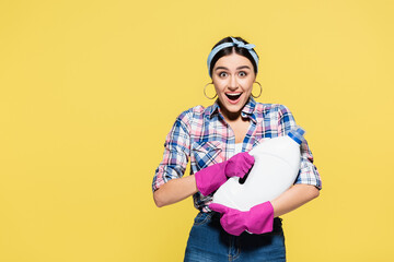 Surprised woman in rubber gloves holding bottle of detergent isolated on yellow