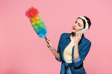 Housewife talking on smartphone and holding dust brush isolated on pink