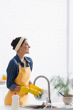 Cheerful woman in apron and rubber gloves washing plate in kitchen