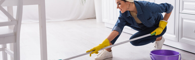 Young smiling woman in rubber gloves washing floor near bucket, banner