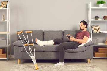 Side view of a man with a broken leg lying on a sofa at home in the living room and browsing social...