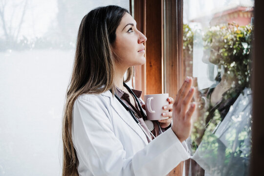 Thoughtful female doctor looking through window while having coffee