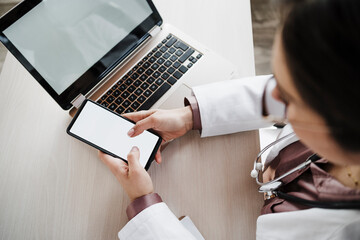 Female doctor using mobile phone by laptop at desk
