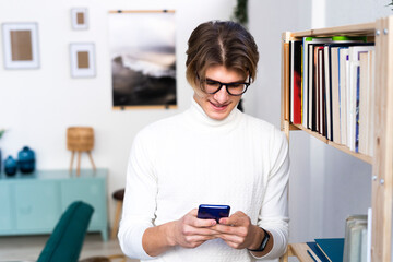 Young man text messaging through smart phone while standing by bookshelf in living room