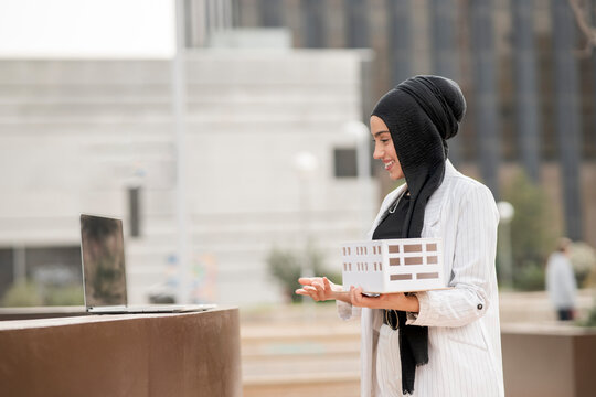 Young female architect holding architectural model during video call on laptop 