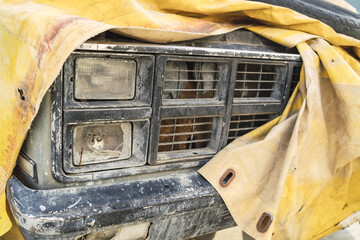 Closeup of the headlight and bumper of an abandoned pickup truck covered with a yellow tarpaulin