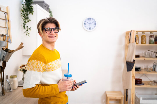 Smiling man wearing eyeglasses holding phone and jar of smoothie in kitchen