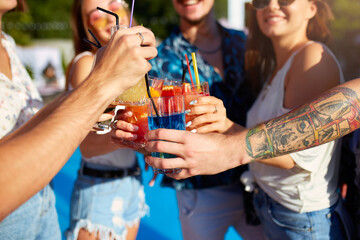 Close view of friends having fun at poolside summer party, clinking glasses with fresh summer cocktails near hotel swimming pool outdoors. People toast drinking juice at luxury villa on vacation.