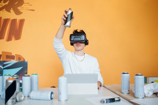 Young man wearing virtual reality simulator while holding spray paint bottle sitting at table in studio