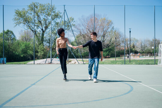 Happy multi ethnic couple walking while holding hands at sports court during sunny day