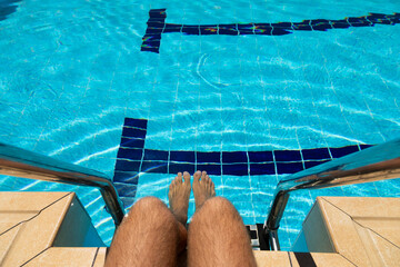 The feet of a young man, sitting on the edge of a pool filled with crystal clear blue water,...