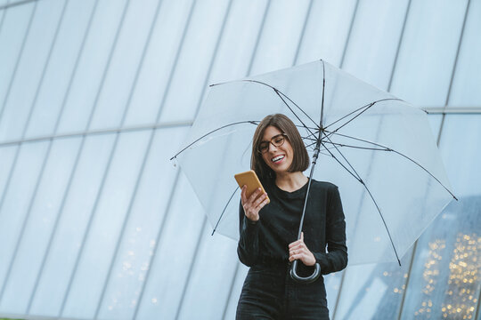 Cheerful young woman with umbrella using smart phone while standing in front of glass building
