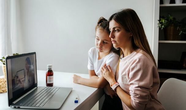 Mother and daughter listening to pediatrician over video call on laptop at home