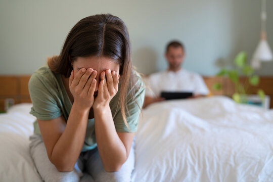 Desperate woman covering her face sitting on bed while boyfriend using digital tablet in background at home