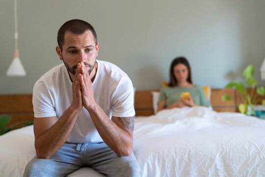 Worried young man sitting on bed while girlfriend using smart phone in background at home