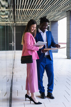 Businesswoman using digital tablet while standing by coworker with file at office corridor