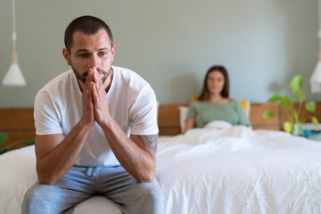 Serious man sitting on bed with girlfriend in background at home