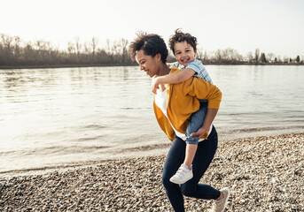 Cheerful woman piggybacking smiling boy while running by lakeshore on sunny day