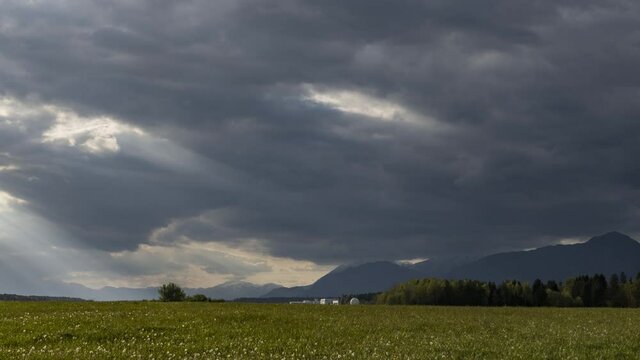 Time lapse of dark clouds moving over green pasture field. Alps mountains in the distance. Sunset in summer season. Right trucking, wide angle