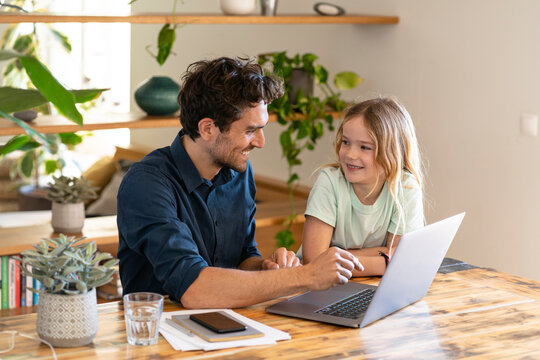 Smiling father and daughter looking at each other in front of laptop at home