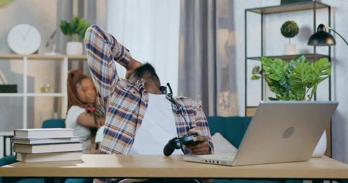 Disappointed african man in headset got defeated during video games on modern laptop. Young father sitting at table and playing on computer while his wife taking care of daughter on background.