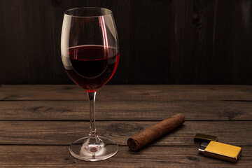 Glass of red wine and cuban cigar with golden lighter on an old wooden table. Focus on the cuban cigar